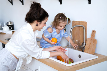 A beautiful young woman with her two year old daughter is washing fruit in the kitchen sink. Fresh bright fruits, apples, oranges, pears. Healthy food for children. The baby helps her mother.