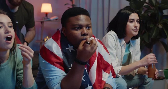Close up of concentrated young people holding different flags while eating and drinking near TV. Multiethnic friends supporting different teams while watching international competition.