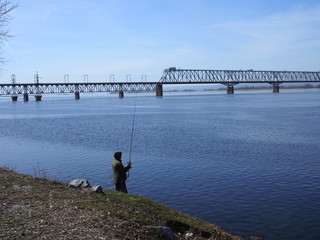 Fototapeta premium young fisherman in a green hoodie sits on the river Bank near the bridge in spring