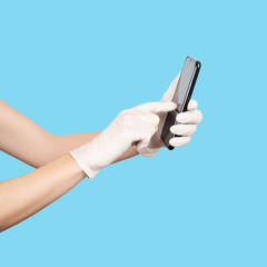 Young woman holding a mobile phone in her hands wearing disposable latex protective gloves isolated on blue.