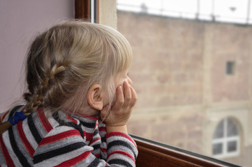 The white-haired blue-eyed girl looks out the window. Quarantined child. The child is sad at the...