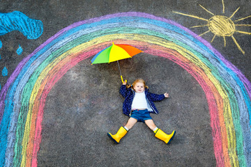 Happy little toddler girl in rubber boots with rainbow painted with colorful chalks on ground...