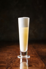 delicious light yellow sour foam cocktail in a long narrow glass and fresh lemon slice, dark wooden background, vertical, side view