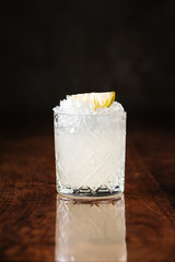 fresh cooling white cocktail with crushed ice and lemon slice in the etched glass on the dark wooden background, side view, vertical