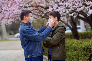 father and son with a face mask are in the city outdoor, blooming trees, spring season, flowering time - concept of allergies and health protection from dusty air
