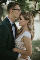 Fototapeta premium Portrait of a lovely bride and groom embrace, kiss and hug each other. Couple in love in the bosom of nature . Handsome groom and beautiful blonde bride posing in the forest. Modern dress and suit