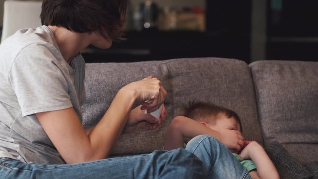 Pretty mother giving some pills to a son laying on a couch. The parent wants to protect the child from coronavirus.