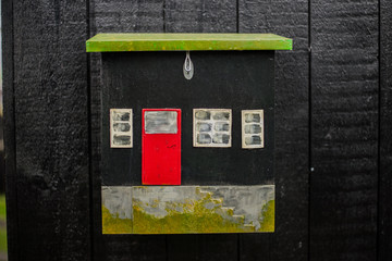 Decorative small black wooden postbox in shape of house with white windows and red door. Black wooden background
