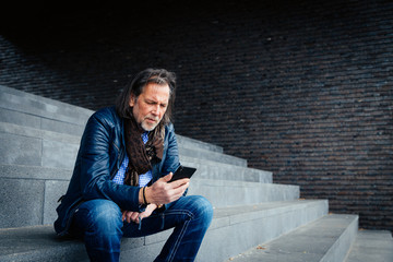 Fototapeta na wymiar Older but cool man with beard and long grey hair uses a mobile phone in an urban environment