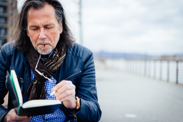 Older but cool man with beard and long grey hair writes in his notebook on the riverbank