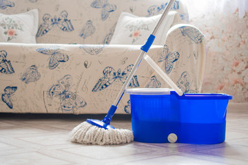 Round MOP and blue bucket on the light linoleum of the house against the background of a soft sofa:...
