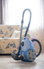 A purple vacuum cleaner stands on the floor against the background of a soft sofa and a window of the house, ready to clean in the morning from dirt and dust, vertically