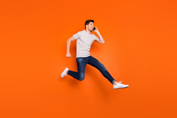 Full length profile photo of funky guy jump high up hold telephone rushing speak friends meeting wear striped t-shirt jeans sneakers isolated bright orange color background