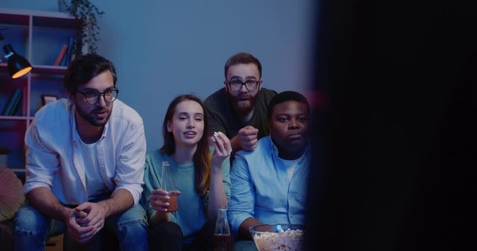 Front view of friends eating popcorn and drinking smiling while watching something . Group of young people talking and point finger at TV while sitting on sofa at home. Concept of leisure.