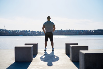 Young sportsman, wearing grey t-shirt, posing in front of blue sky during sunset. Muscleman standing on concrete platform by the lake, showing his power.