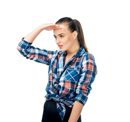 Young female keeps hand near forehead. Dark hair combed in knot. Dressed in casual shirt. Notices something in distance. Poses against white background.