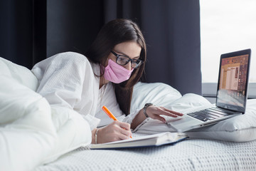 Obraz premium Quarantine, a woman in glasses and a mask lies in bed working on a laptop online. Stay at home.