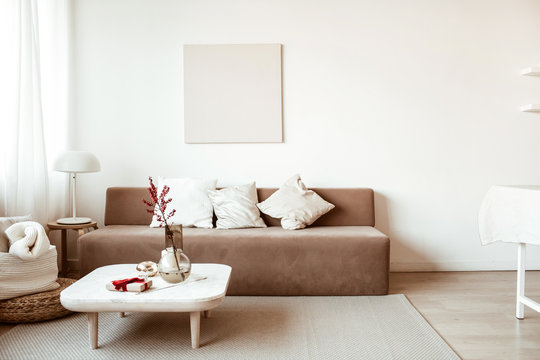 Modern interior design. Stylish bright living room decorated with comfortable sofa, coffee table, flowers, painting, white walls. Minimalistic apartment for rent.