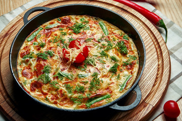 Appetizing Spanish fritata omelet baked in a pan with vegetables, meat and melted cheese on a wooden background. Close up view.
