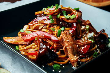 Udon noodles with baby corn, bell pepper, onions, carrots and veal fried on a wok with teriyaki sauce in black plate on wooden background. Tasty asian street food