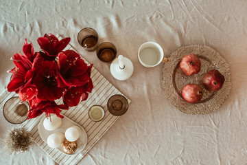 Decorated kitchen table with pomegranate on golden plate, red flowers bouquet, kitchenware, candles on tan beige table cloth.