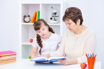 A girl with down syndrome is reading a book with her teacher at home at the table. Equal opportunity for disabled concept