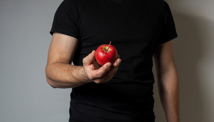 A man in a black T-shirt offers a red apple on a gray wall background