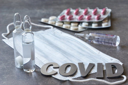 Metal Lettering Covid And Three Vials Of Medicine. Medical Mask, Tablets On A Grey Textured Background