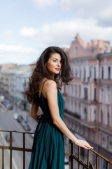 Fototapeta premium Girl with long black hair in a green dress stands on a city balcony