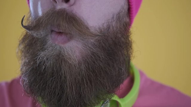 Close-up of tattooed Caucasian man with beard and mustache in pink t-shirt and hat eating a Chupa Chups,sweet candy,looking at camera,with headphones isolated on yellow background