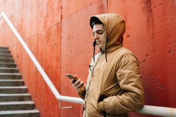 a young guy in a brown jacket on the street, standing near a red wall uses his smartphone . street style