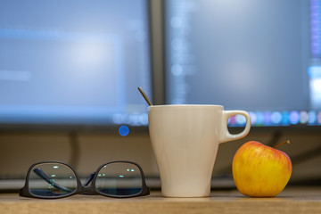 White cup of coffee or tea, glasses and a red apple on blurred background of personal computer screen.