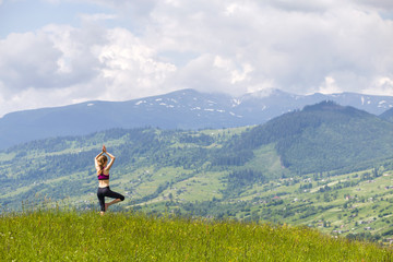 Fototapeta na wymiar Attractive slim young woman doing yoga exercises outdoors on background of green mountains on sunny summer day.
