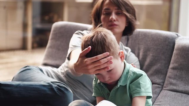 Mother and son are laying on a sofa and mother is touching the forehead of the son. She wants to check the temperature of her kid and puts on a disposable mask.