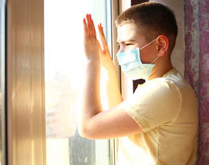 Portrait of a Caucasian boy in a medical mask stands and looking out the window. Misses you at home. Quarantine protection against coronavirus. Home insulation