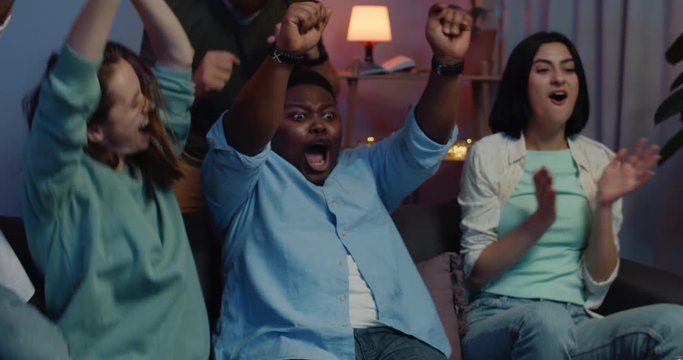 Excited friends rejoicing, raising hands while supporting team in sport match. Group of young people having good time while sitting on sofa at home. Concept of leisure.