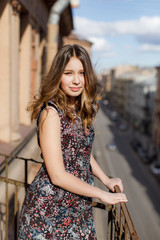 Fototapeta premium Young girl in a floral dress stands on a city balcony