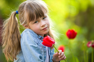 Cute pretty smiling child girl with gray eyes and long hair with bright red tulip flower on blurred sunny green bokeh background. Love to nature concept.