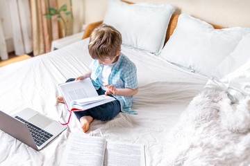 Distance learning online education. Caucasian boy with book. and pen studying at home and doing school homework. Sitting on bed with training books.