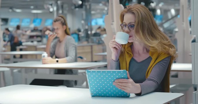 Adult caucasian businesswoman surfing internet using tablet PC enjoying morning coffee sitting at city cafe food court. Attractive cute modern woman using social networks chatting