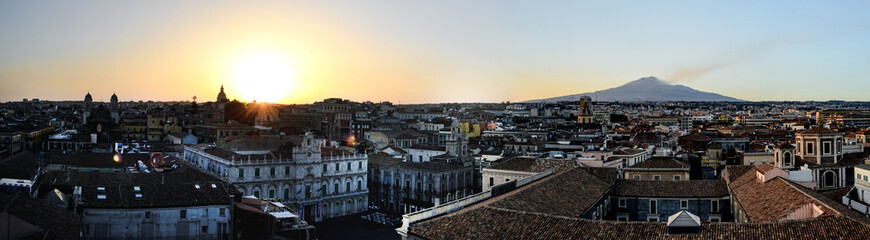 Naklejka na ściany i meble Catania, Sicily in Italy. Aerial view of the city roofs at sunset with the incredible Etna vulcano smoking in the background, nice warm colors and soft light.