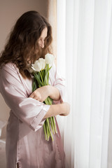 Beautiful young girl staying near a window with tulips bouquet