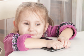 Fototapeta premium happy smiling three year old girl in a purple dress sits at a table and plays with a tablet