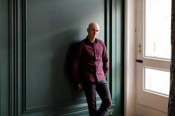 Bald man in a plaid shirt and gray jeans stands near a window against a green wall