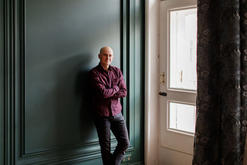 Bald man in a plaid shirt and gray jeans stands near a window against a green wall