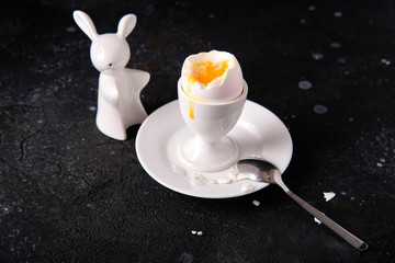 Fototapeta na wymiar Boiled egg in a ceramic white stand on a white saucer with a spoon and ceramic salted in the form of a rabbit on a dark table. Breakfast.