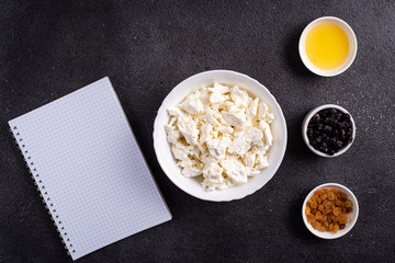 Cottage cheese in a white plate, separately honey, raisins, next to a white notebook on a black background. Photography for recipes.