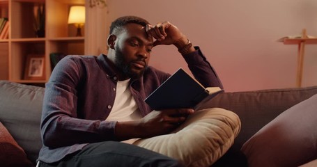 Handsome african guy leaning head with hand while sitting on sofa at living room. Young man spending free time while reading interesting book. Concept of leisure and hobby. - Powered by Adobe