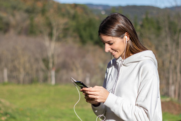 Pretty smiling young woman breathes fresh air in the park listening to music on headphones and holding a smartphone. She is wearing a light sweatshirt and has long brown hair.