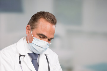 Tired male doctor in lab coat and tie in front of a clinic room